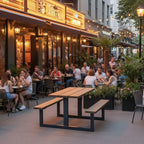 Outdoor patio seating at a restaurant with multiple people dining at tables, potted plants, and wooden picnic-style tables on a city sidewalk during the evening.