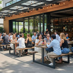 Outdoor dining area at a modern restaurant with multiple groups of people seated at long wooden tables under a glass canopy with string lights. The setting includes large windows, a wood-and-black facade, and potted plants.