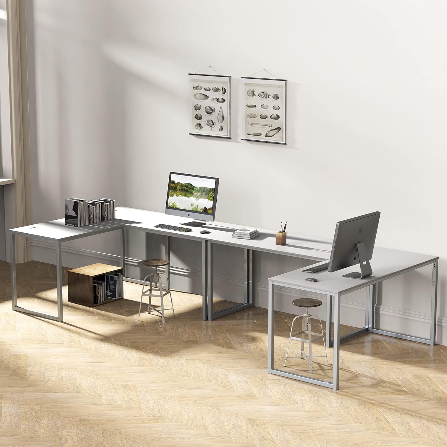 Modern L-shaped office desk with metal frame, stools, computers, and wall art in a bright workspace