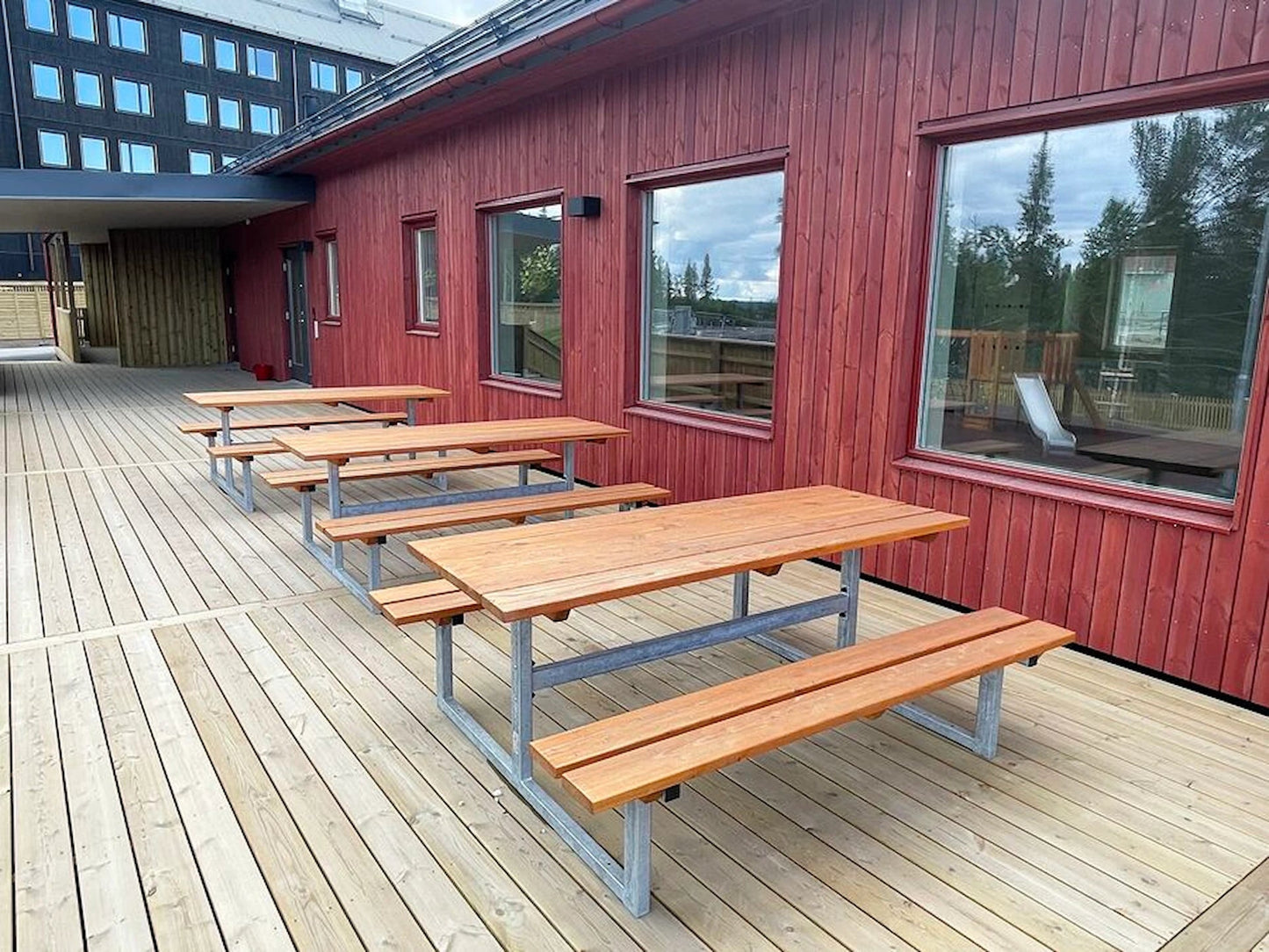 Wooden outdoor bench tables with metal frames on a wooden deck by a red building