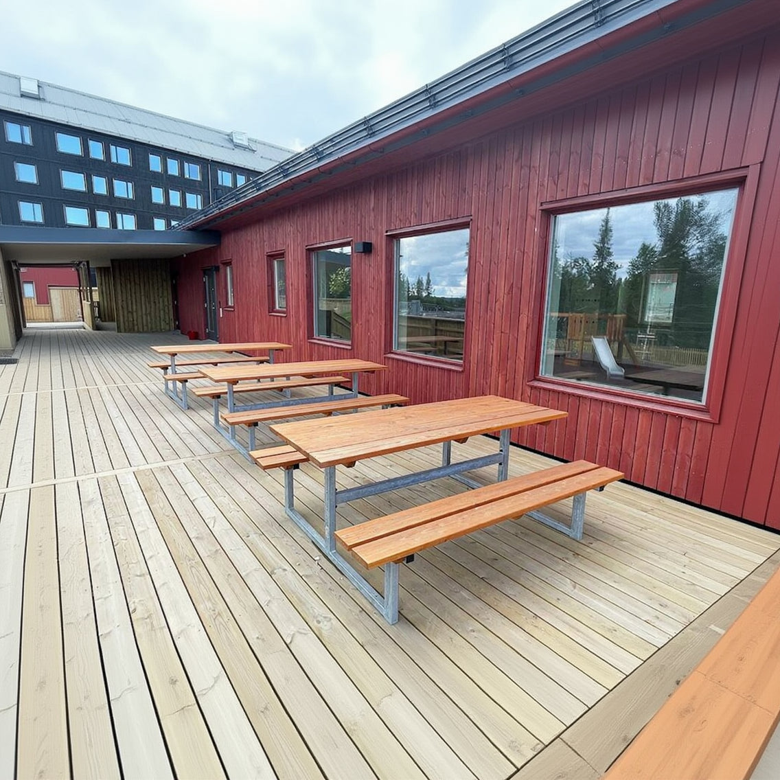 Wooden outdoor bench tables with metal frames on a wooden deck by a red building