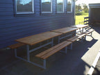 Outdoor wooden bench and table set with metal frame on paved area beside blue building