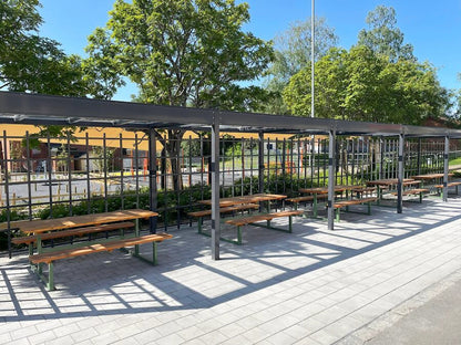 Outdoor wooden benches and tables under metal pergolas on a paved patio, surrounded by trees