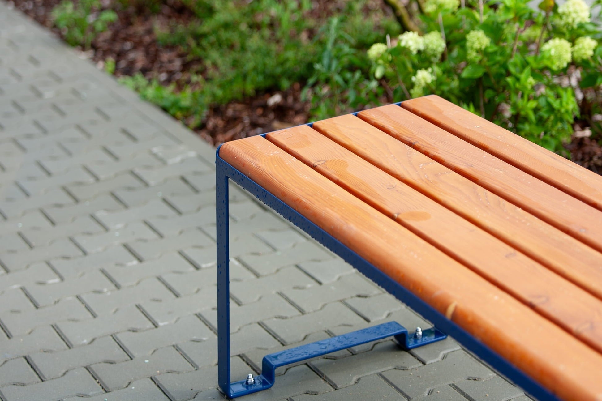 Backless outdoor wooden bench with blue metal frame on a paved walkway near greenery