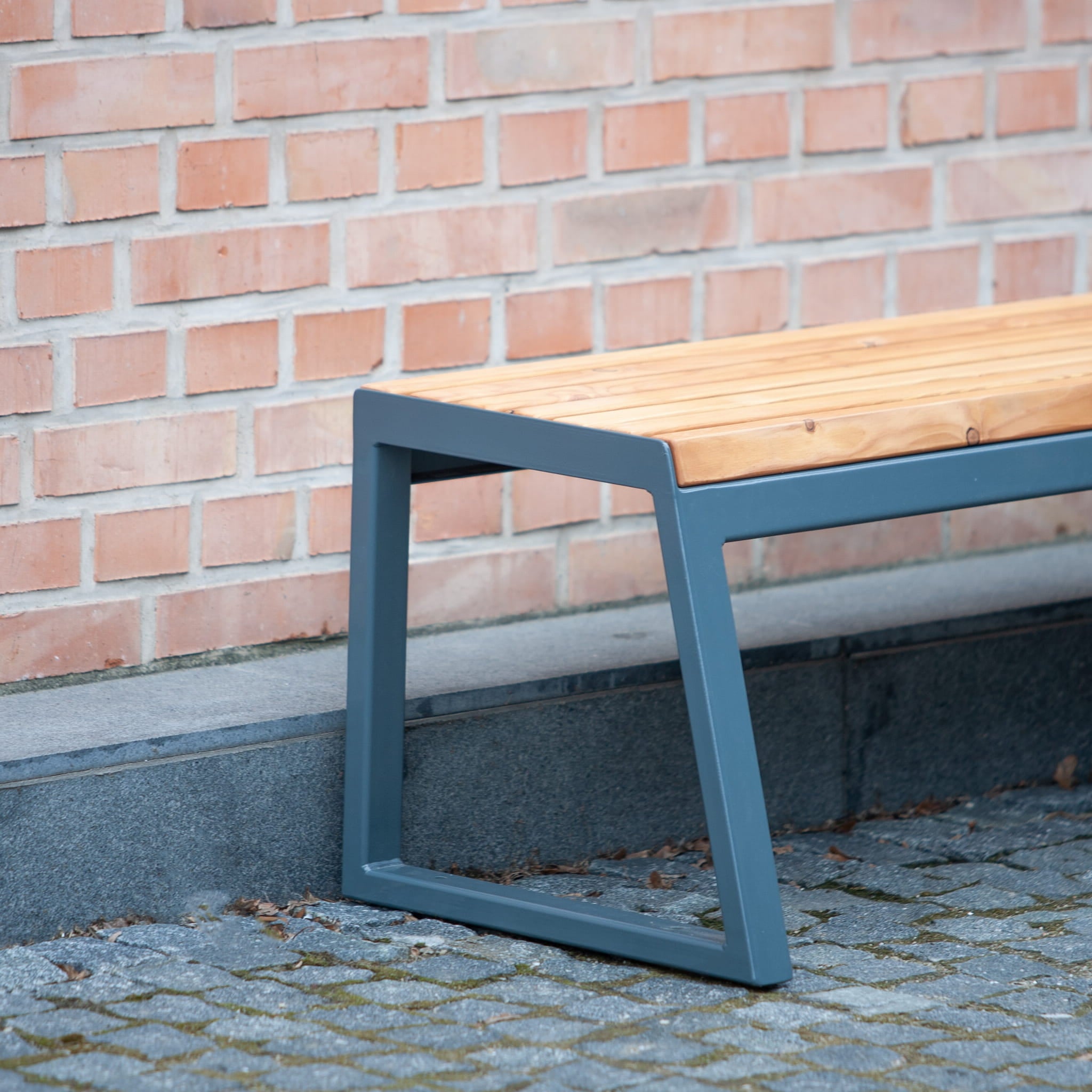 Modern backless bench with wooden seat and metal frame, placed outdoors by a brick wall