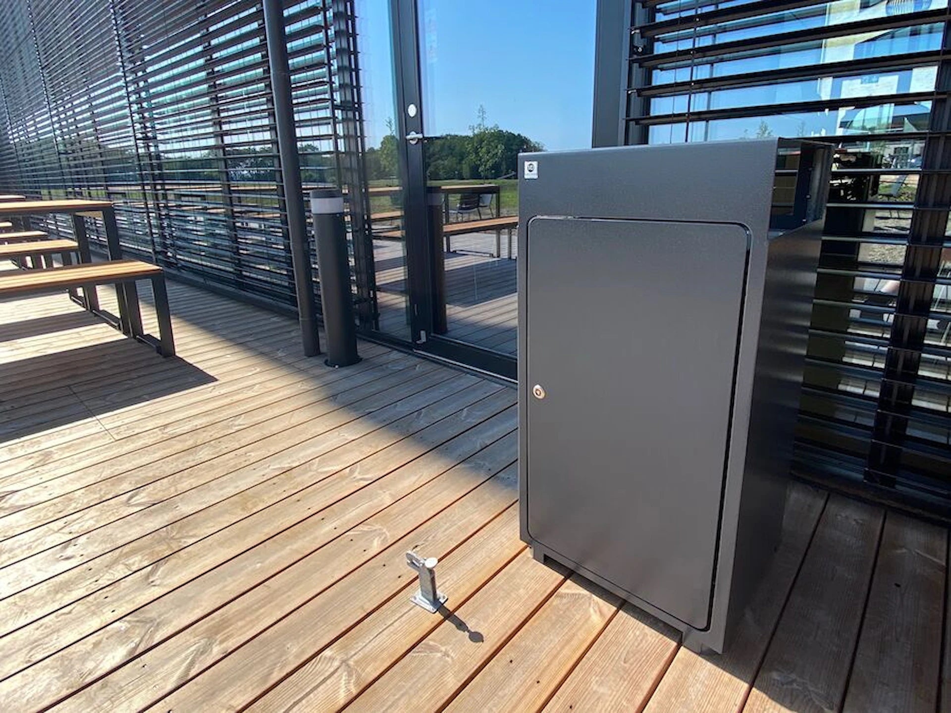 Modern outdoor metal wastebasket on wooden deck near benches and glass wall
