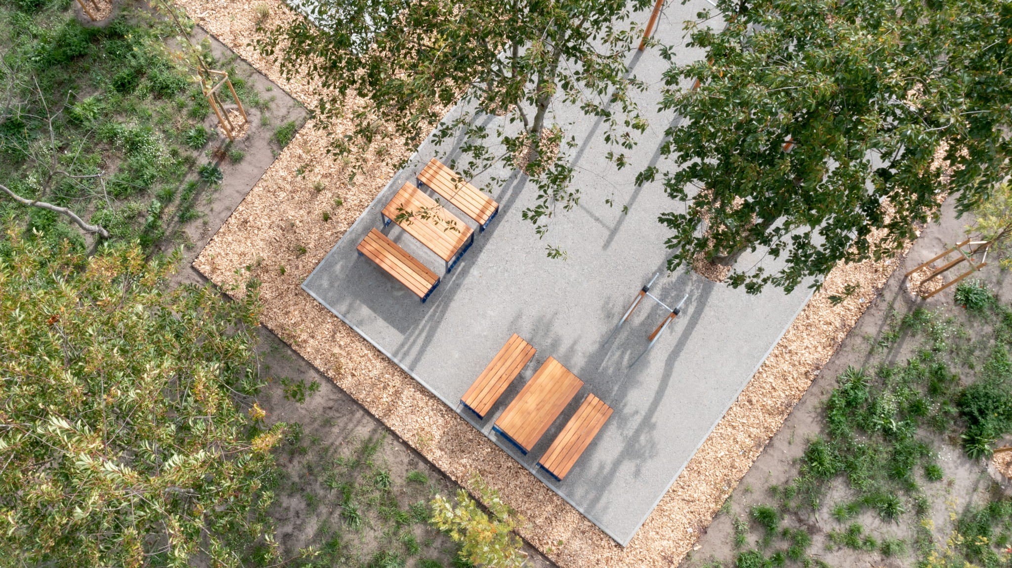Outdoor rectangular wood tables with benches, seating 4-6, on a paved patio surrounded by trees.