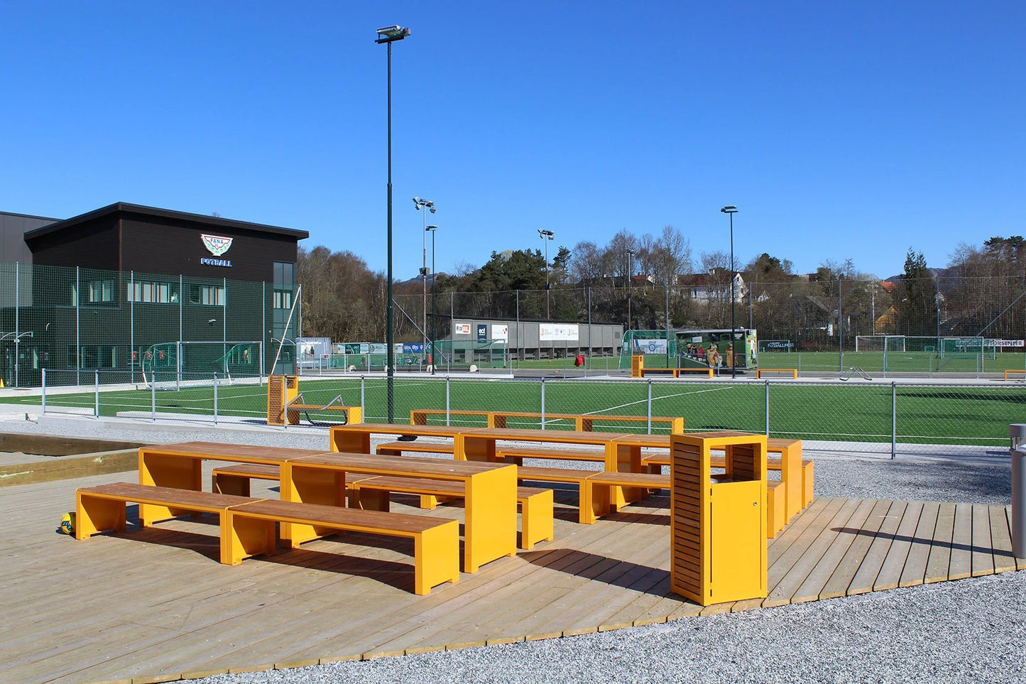 Yellow outdoor wastebasket next to modern wooden benches on a patio by a sports field