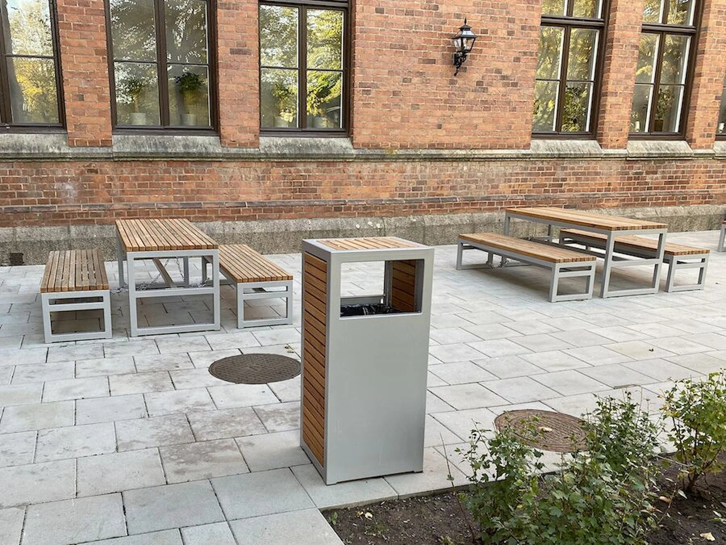 Modern outdoor wastebasket with metal frame and wood panels, surrounded by matching benches and tables on a paved patio.