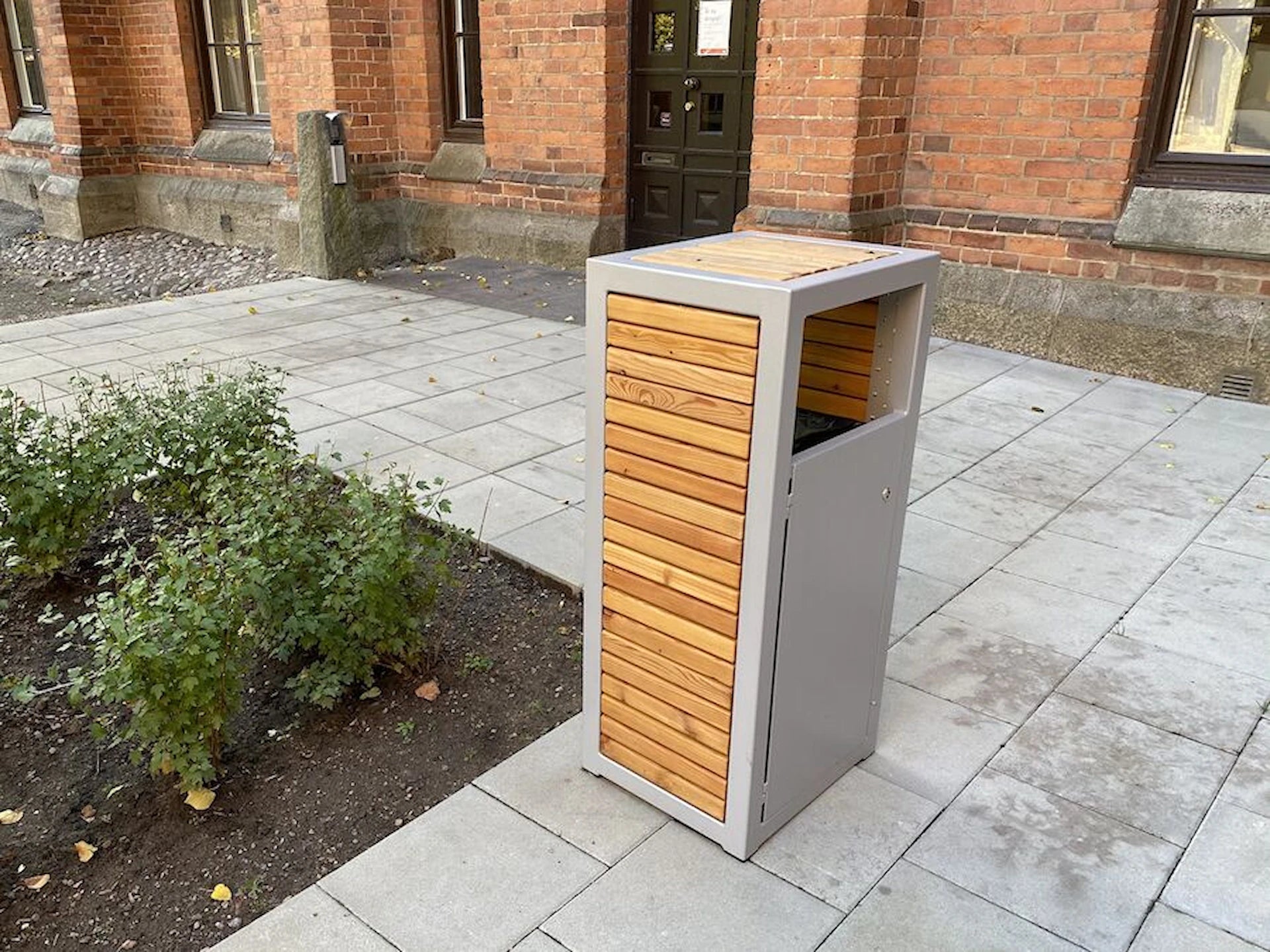 Modern outdoor wastebasket with wood panel design on a paved area near brick building